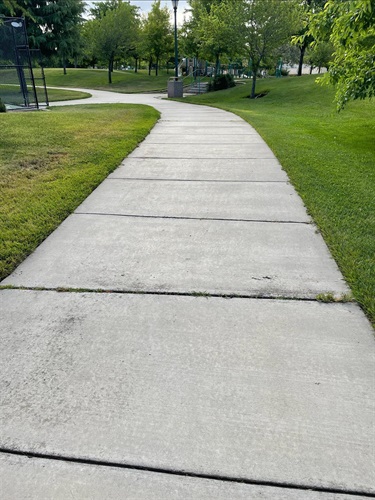 Wide concrete walking path curving through a grassy park, bordered by mature trees, landscaped areas, and open green space.