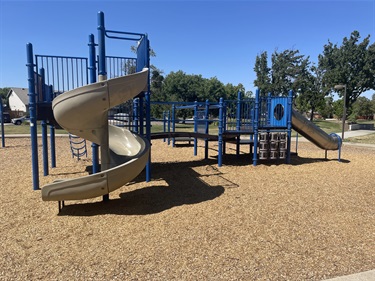 Playground with a spiral slide, straight slide, climbing structures, and activity panels on a wood chip surface, surrounded by open grassy areas and mature trees in a community park.