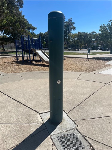 Dark green outdoor drinking fountain mounted on a concrete pad beside a playground, with walking paths, play equipment, and mature trees visible in a sunny community park.