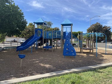 Blue playground structure with a slide, tunnels, and a spinning seat, on a mulch surface with a paved path and parked cars in the background.