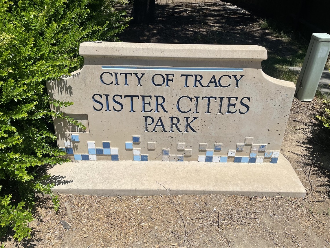 Stone entrance sign reading 'City of Tracy, Sister Cities Park' with a blue tile mosaic border, flanked by a shrub and a white post.