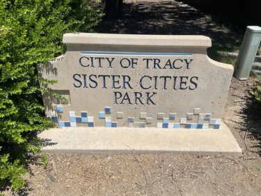 Stone entrance sign reading 'City of Tracy, Sister Cities Park' with a blue tile mosaic border, flanked by a shrub and a white post.