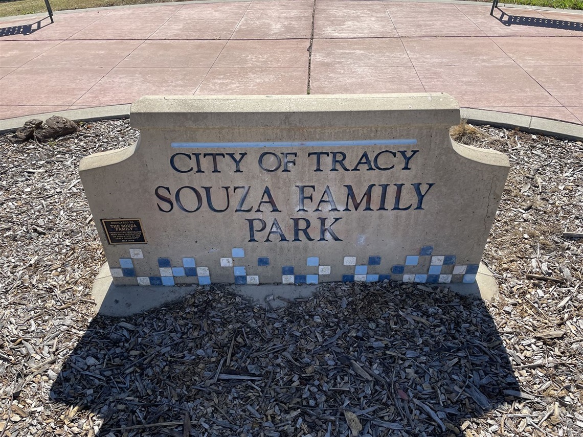 Stone entrance sign reading 'City of Tracy, Souza Family Park' with a blue tile mosaic border, set against a gravel background.