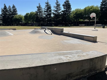 Concrete skate park with ramps, rails, and bowls, surrounded by trees under a partly cloudy sky.