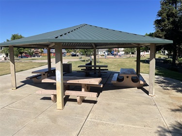 Covered picnic shelter with multiple picnic tables, a barbecue grill, and concrete seating area in an open park with grassy fields and nearby trees.