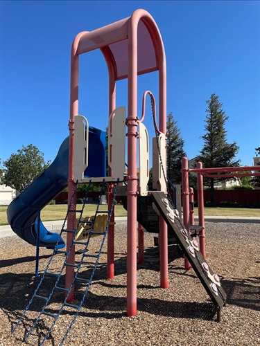 Playground tower with a curved blue slide, rope climbing net, and inclined climbing wall, connected to a larger play structure on a wood chip safety surface.