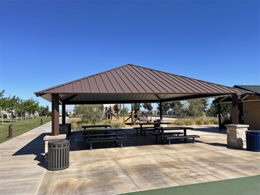 Covered picnic pavilion with multiple picnic tables on a concrete pad, surrounded by walking paths, open grassy areas, landscaped plantings, and nearby park amenities in a community park under a clear blue sky.