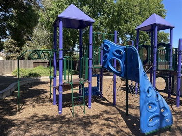 Purple and blue playground climbing structure with a slide and rope netting, on a mulch surface with a fence and trees in the background.