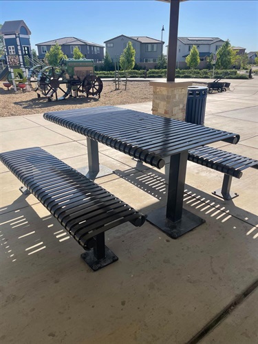 Covered picnic shelter with a metal picnic table and benches on a concrete pad, overlooking a playground with play equipment, decorative wagon-themed play features, walking paths, and nearby park amenities in a community park.