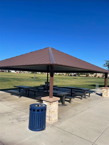 Covered picnic pavilion with multiple picnic tables on a concrete pad, surrounded by open grassy areas, walking paths, and park amenities, with a nearby trash receptacle under a clear blue sky.