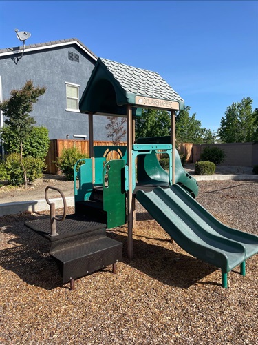 Green and tan playground structure with a peaked roof, slide, and spring rocker, on a mulch surface with a house and fence in the background.
