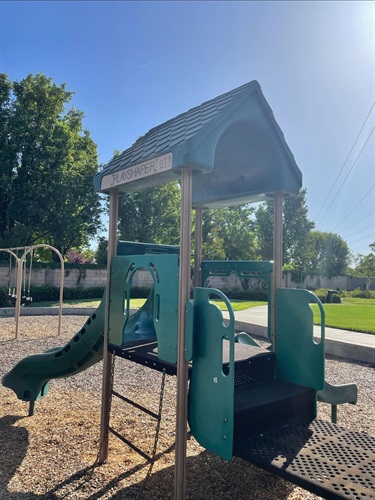 Small playground structure with a peaked roof, slide, and climbing steps, on a mulch surface with a fence and trees in the background.