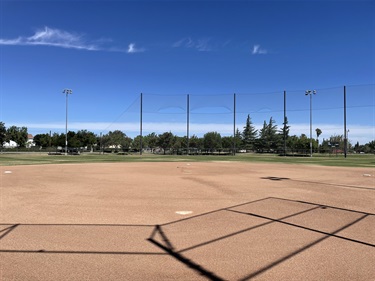 Baseball field with a dirt infield, home plate, backstop fencing, tall ball stop netting, and field lighting, viewed across the playing surface in a community sports park under a clear blue sky.