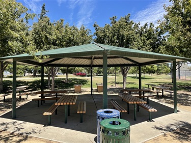 Large covered picnic pavilion with multiple picnic tables on a concrete pad, surrounded by mature shade trees, open grassy areas, and park amenities, with nearby trash and recycling receptacles in a community park.
