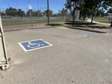 Accessible parking spaces with painted wheelchair symbols in a park parking lot, adjacent to a chain-link fence enclosing sports fields and surrounded by mature trees under a clear blue sky.