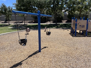 Two-bay swing set with bucket swings for young children on a wood chip safety surface, adjacent to an accessible playground structure with ramps and activity panels, surrounded by open grassy areas and mature shade trees in a community park.