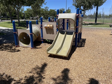 Playground with a double slide, crawl tunnel, activity panels, and elevated play structure on a wood chip safety surface, surrounded by open grassy areas, mature shade trees, and walking paths in a community park.