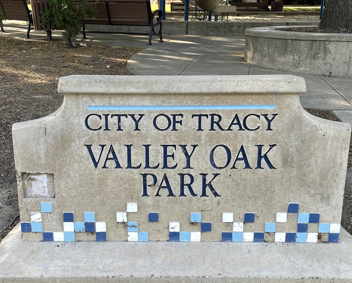 Stone entrance sign reading 'City of Tracy, Valley Oak Park' with a blue tile mosaic border, framed by trees.