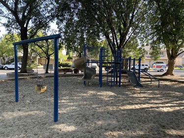 Playground swing set and climbing structure on a sand surface, with a car parked nearby and trees in the background.