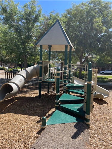 Playground with climbing structures, elevated platforms, a tube slide, and an open slide on a wood fiber safety surface, surrounded by mature trees and open park space.