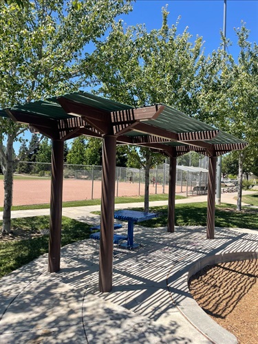 Wooden pergola with shaded bench seating along a concrete walkway, overlooking a baseball field, and surrounded by mature shade trees, landscaped park areas, and open green space in a community park.
