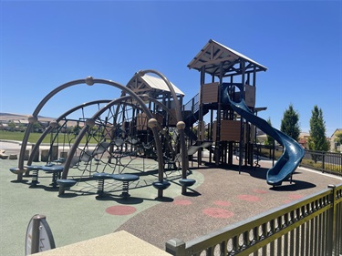 Large playground with a multi-level play structure, spiral slide, rope climbing bridge, climbing nets, and balance stepping pods on a rubberized safety surface, surrounded by open grassy areas, nearby residential homes, and walking paths in a community park under a clear blue sky.