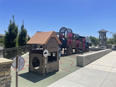 Playground with a playhouse, interconnected play structures, tunnels, climbing features, and activity panels on a rubberized safety surface, bordered by a low concrete seating wall and concrete walkways, with a gazebo visible in the background in a community park.