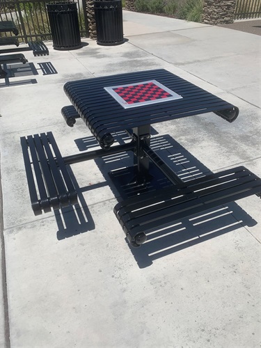 Accessible game table with an integrated checkerboard playing surface and attached benches on a concrete plaza, surrounded by nearby benches, trash receptacles, landscaped areas, and walking paths in a community park.