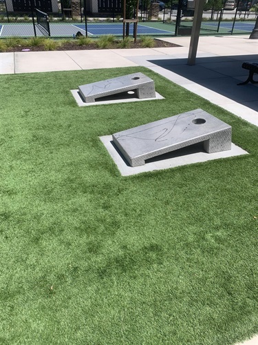 Two cornhole boards installed on a rectangular artificial turf play area, located beside a covered picnic pavilion and adjacent to fenced sports courts, with concrete walkways and landscaped park amenities in a community park.
