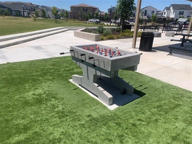 Accessible outdoor foosball table on an artificial turf play area, surrounded by concrete walkways, benches, trash receptacles, landscaped areas, and open grassy space in a community park.