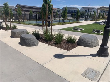 Concrete walkways surrounding a landscaped park plaza with decorative boulders, ornamental shrubs, a decorative light pole, cornhole boards on an artificial turf play area, and fenced pickleball courts, with nearby residential homes in the background under a clear blue sky.