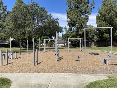 Outdoor fitness area with pull-up bars, balance beams, parallel bars, and other exercise stations on a wood chip surface, surrounded by mature trees and open grassy areas in a community park.
