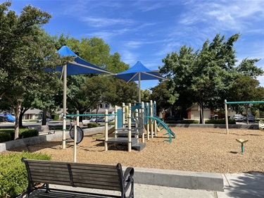 Playground with slides, climbing structures, and shade canopies on a wood chip surface, surrounded by benches, mature trees, and landscaped park areas in a community park.