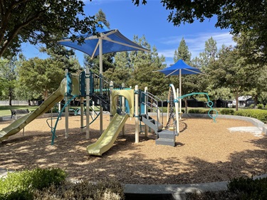 Playground with slides, climbing structures, activity panels, and shade canopies on a wood chip surface, surrounded by mature trees and landscaped park areas in a community park.