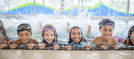 Children smiling at the edge of a swimming pool wearing goggles, with water splashing behind them.