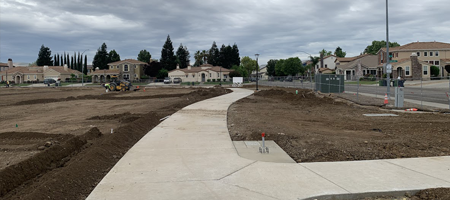 “New concrete walkway under construction through a park area, with dirt landscaping, equipment, and homes visible in the background.