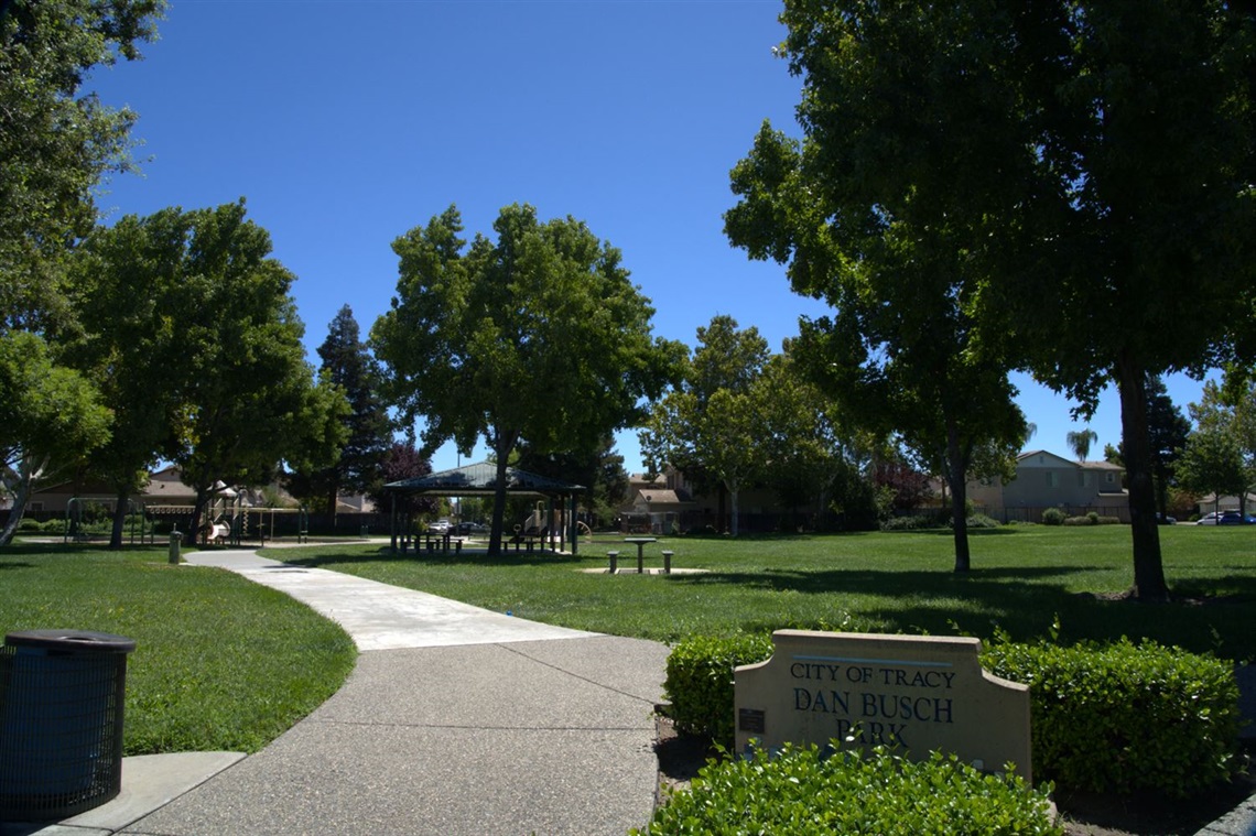 A paved walking path curving through a grassy park area, lined with trees and a low wall, with hills visible in the distance.