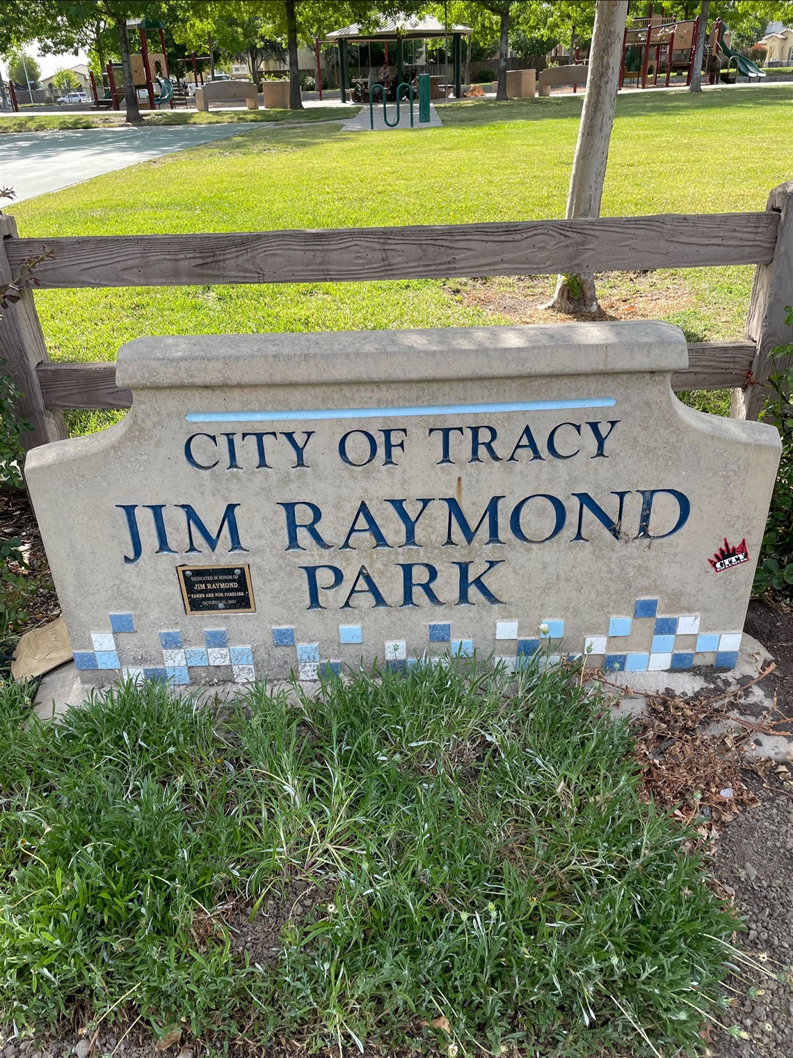 Stone entrance sign reading 'City of Tracy, Jim Raymond Park' with a blue tile mosaic border, in front of a wooden fence, lawn, and playground equipment.