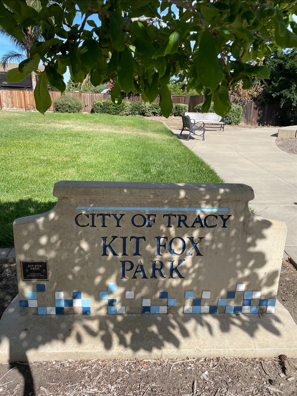 Stone entrance sign reading 'City of Tracy, Kit Fox Park' with a blue tile mosaic border, in front of a grassy lawn, bench, and trees.