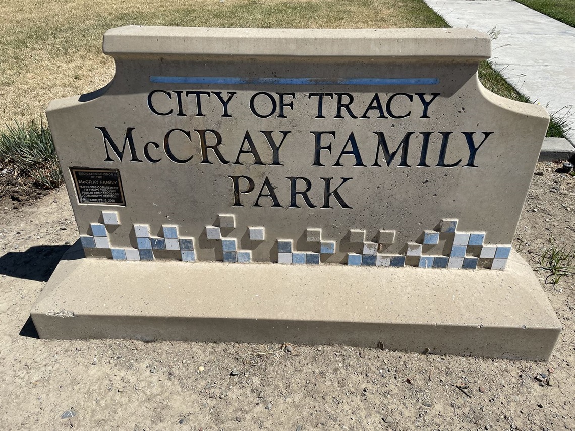 Stone entrance sign reading 'City of Tracy, McCray Family Park' with a blue tile mosaic border, set against a plain gravel background.