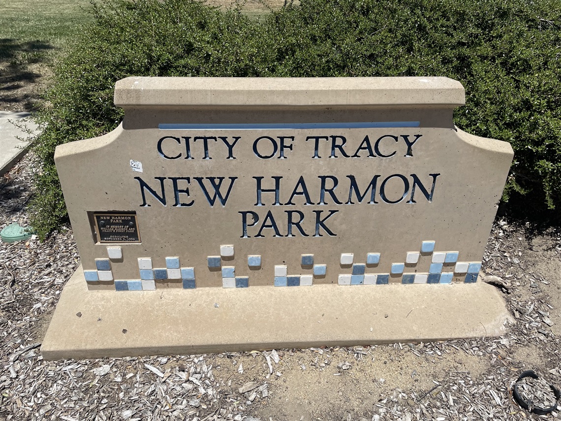 Stone entrance sign reading 'City of Tracy, New Harmon Park' with a blue tile mosaic border, set against a gravel and shrub background.