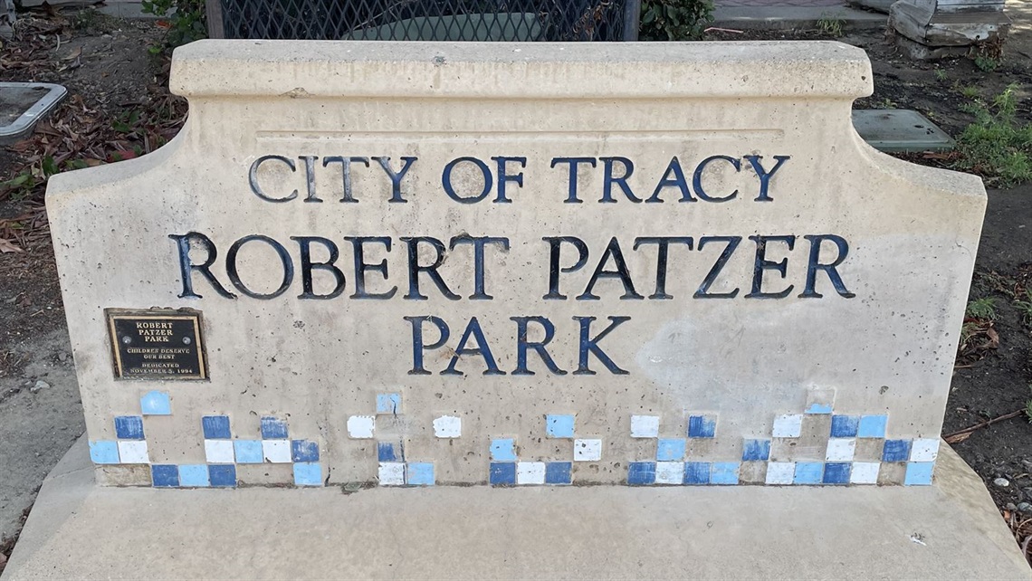 Stone entrance sign reading 'City of Tracy, Robert Patzer Park' with a blue and tan tile mosaic border, framed by greenery.