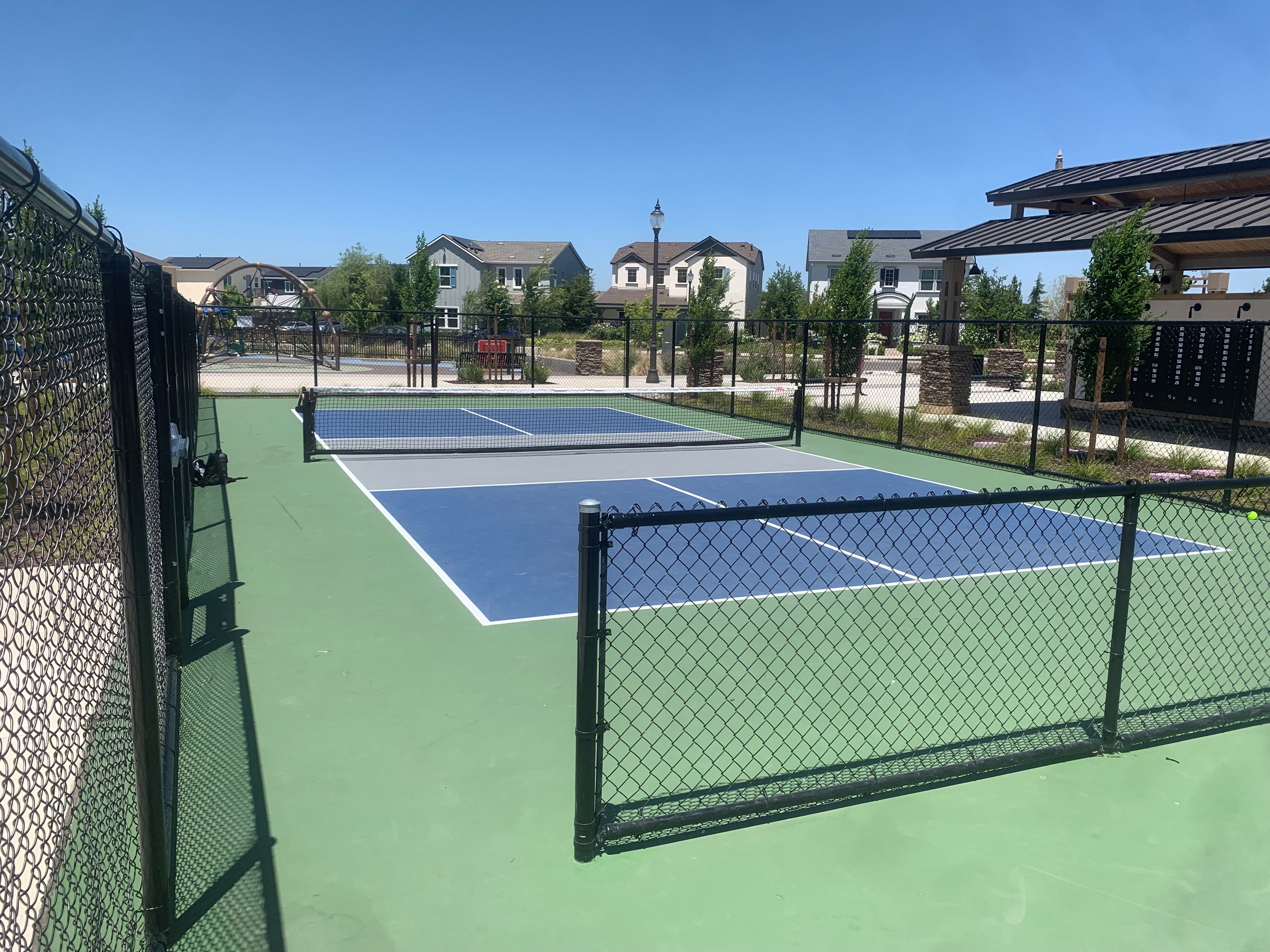 Fenced outdoor pickleball courts with blue and green playing surfaces, surrounded by concrete walkways, landscaped planting beds, nearby residential homes, and a covered park pavilion in a community park under a clear blue sky.