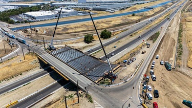 Aerial view of a bridge construction project spanning a major roadway interchange, with cranes lifting materials and crews working on the bridge deck above active traffic lanes.