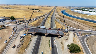 Aerial view of a bridge construction project crossing a divided highway, with cranes and construction crews working on the bridge deck above active traffic lanes in a dry, open landscape.