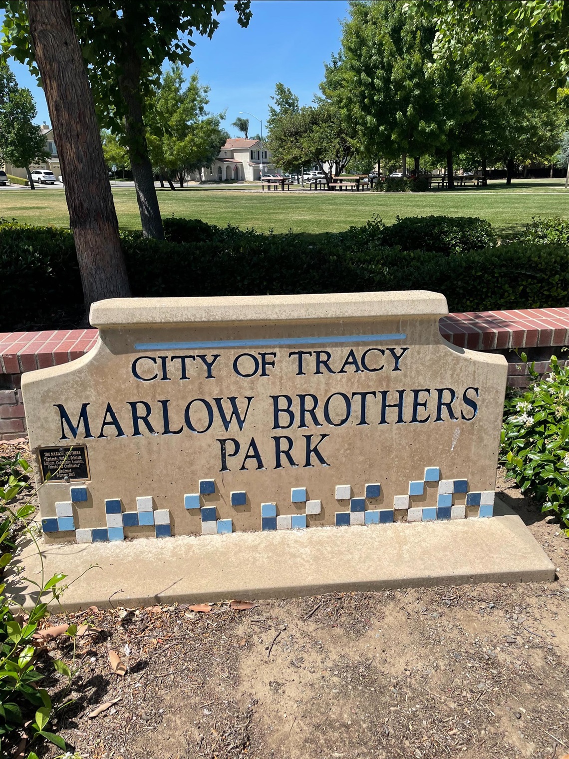 Stone entrance sign reading 'City of Tracy, Marlow Brothers Park' with a blue tile mosaic border, in front of a lawn, trees, and a distant gazebo.
