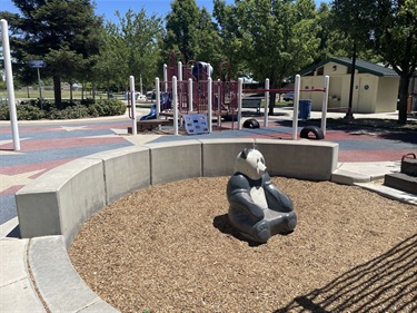 Playground featuring a panda sculpture on a wood chip safety surface, surrounded by a low curved concrete seating wall, with play structures, rubberized play surfacing, and mature shade trees in a community park.