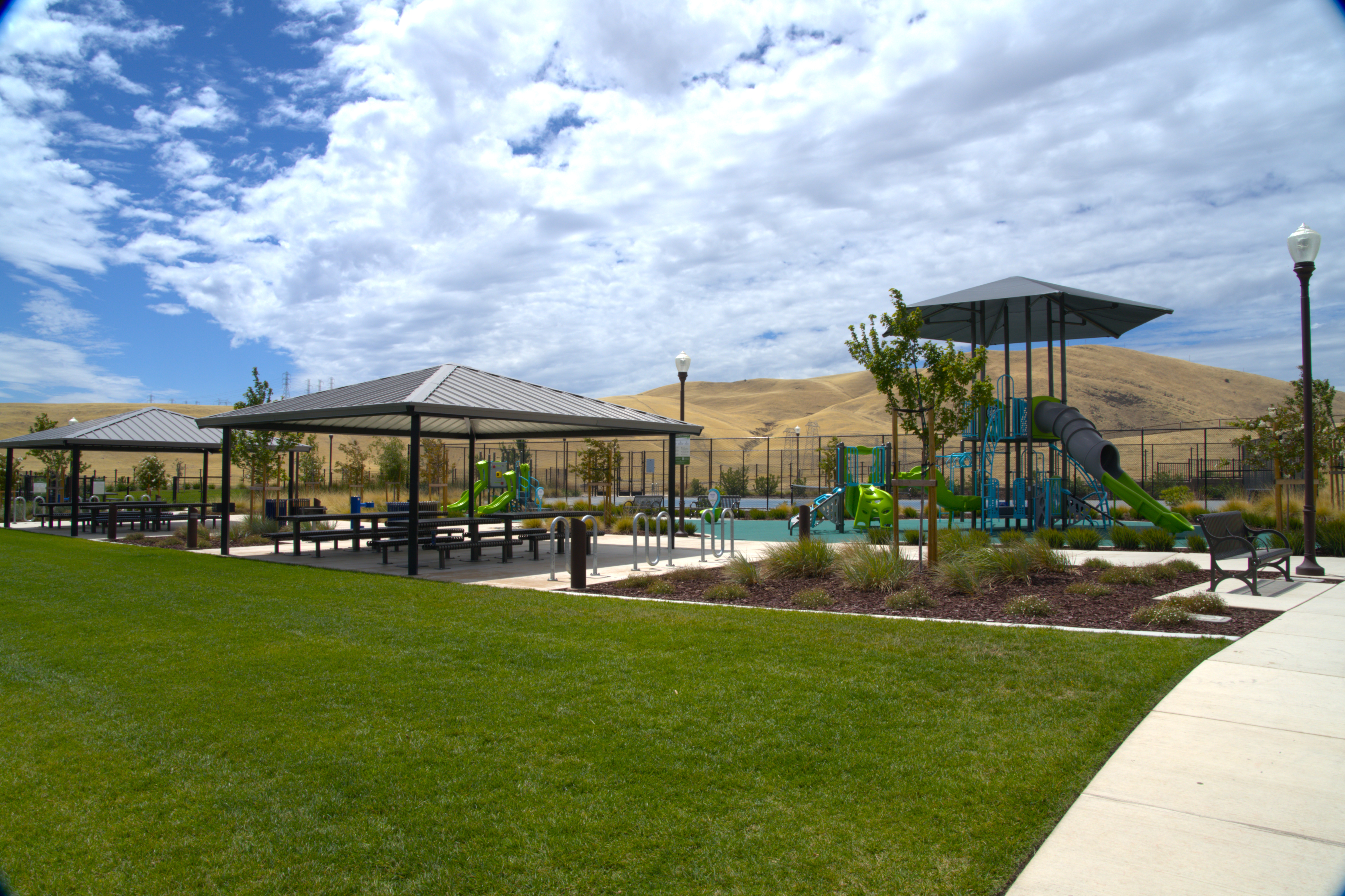 A park with a shaded pavilion, a colorful play structure, and a green lawn in the foreground, set against a hillside backdrop.