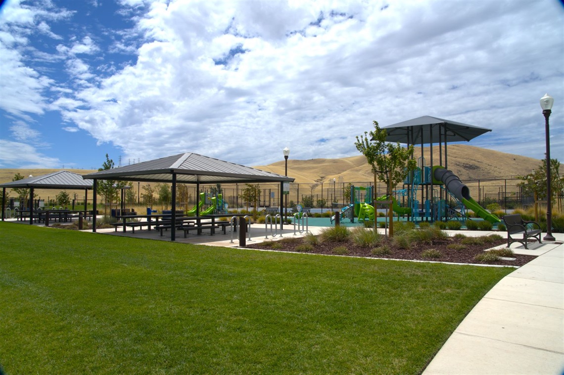 A park with a shaded pavilion, a colorful play structure, and a green lawn in the foreground, set against a hillside backdrop.