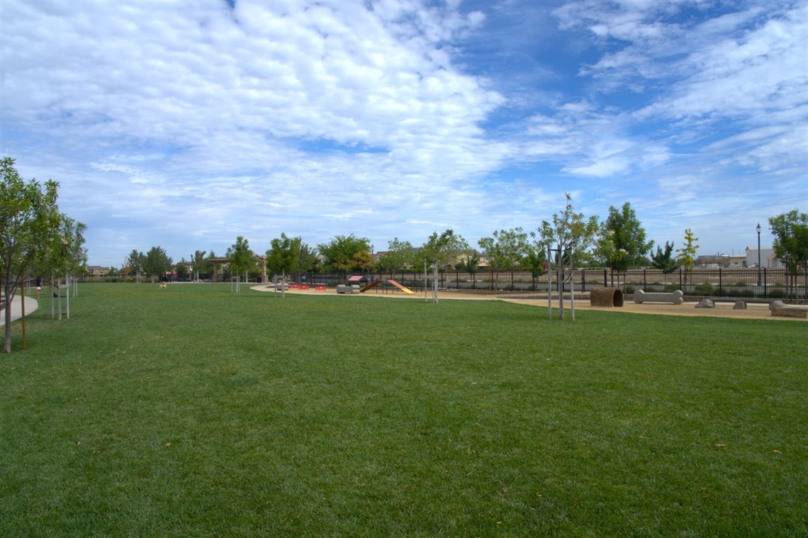 A large open grass field bordered by a low fence, with playground equipment, trees, and houses visible in the background under a partly cloudy sky.